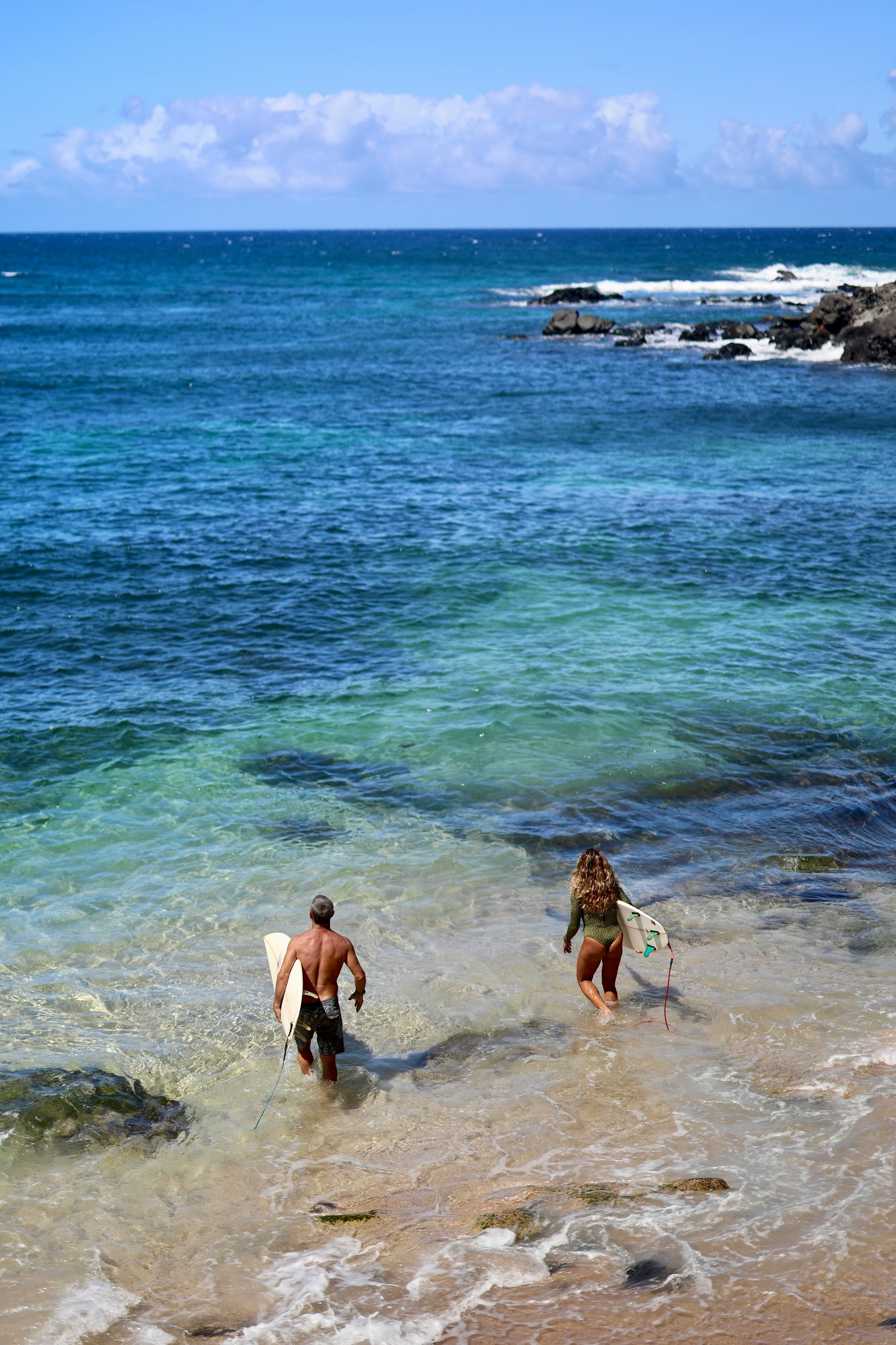Two surfers with boards walk into clear blue ocean water from a sandy beach under a bright sky.